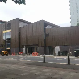 Budynek w trakcie budowy z brązową elewacją panelową, widoczny wózek widłowy i elementy zabezpieczające teren budowy, na tle szarego nieba i wysokiego budynku mieszkalnego.