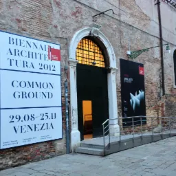 Wejście do budynku z cegły z plakatem Biennale Architektury 2012 i rampą dla niepełnosprawnych.