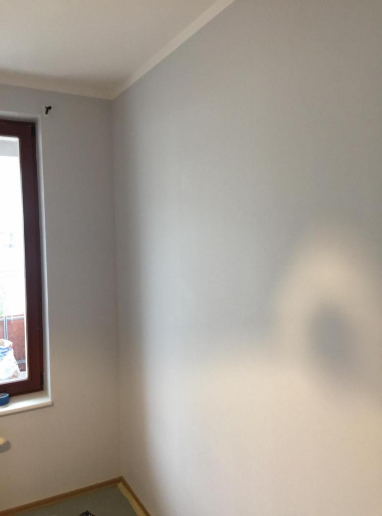 Świeżo pomalowane na szaro ściany w rogu pokoju, widoczne okno z brązową ramą i parapetem, listwa przypodłogowa zabezpieczona folią malarską.