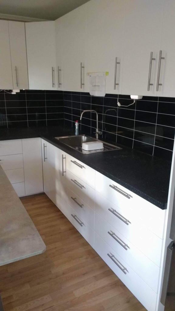 Białe szafki kuchenne z czarnym blatem i czarnymi płytkami na ścianie, widoczny zlew z baterią, drewniana podłoga.