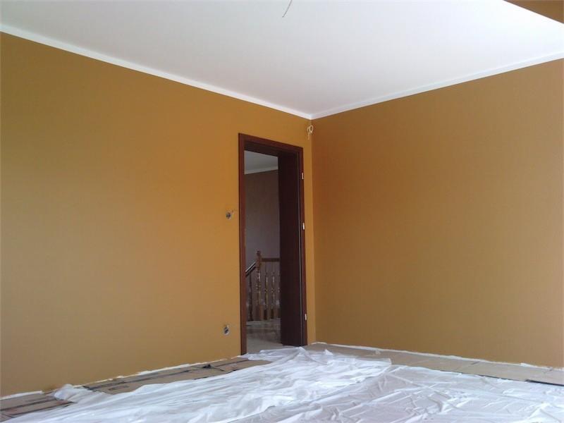 Pomarańczowe ściany świeżo pomalowanego pokoju z białym sufitem i listwami, zabezpieczona podłoga folią malarską, widoczne drzwi do kolejnego pomieszczenia ze schodami.