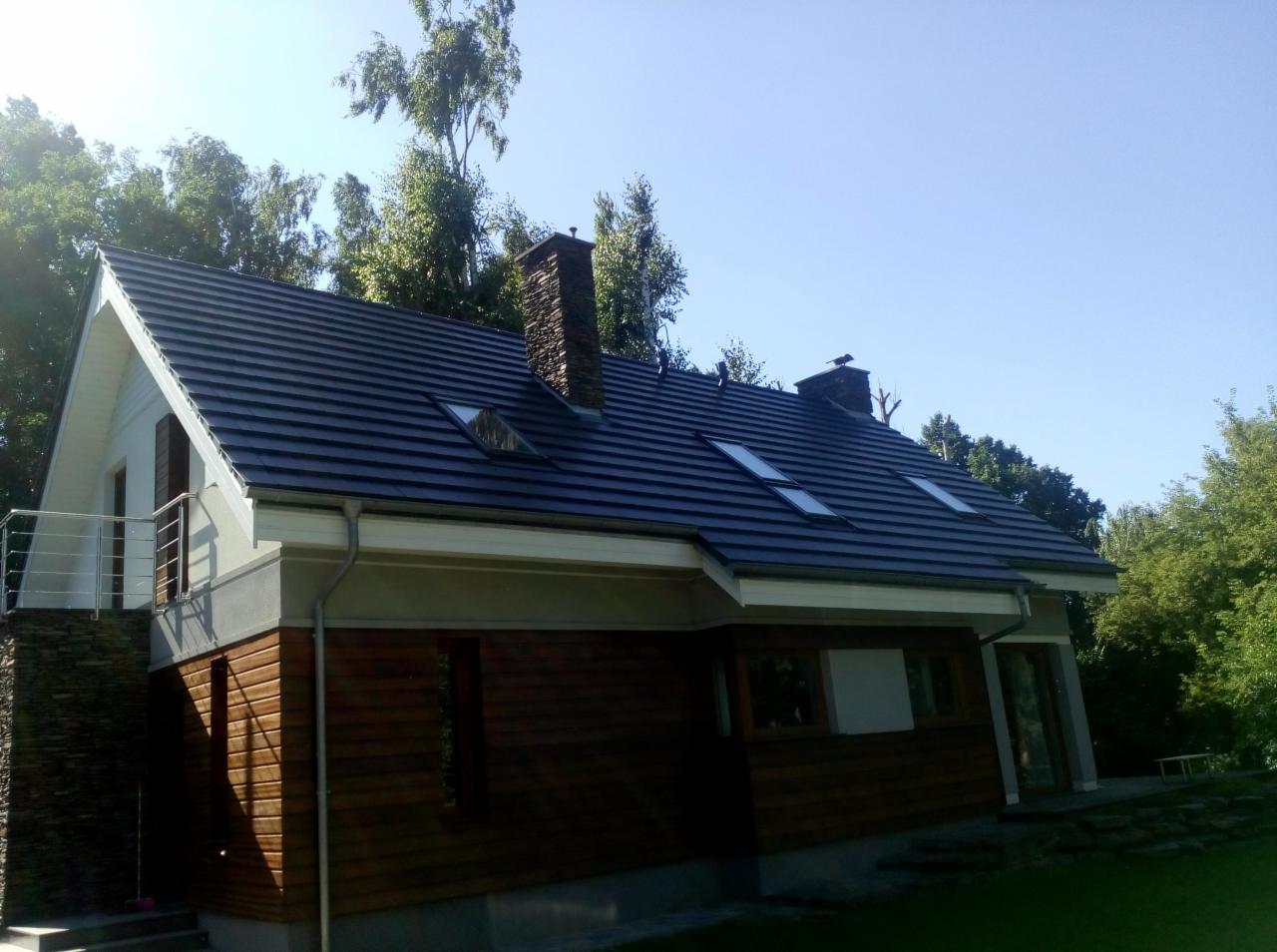 Dom jednorodzinny z ciemną dachówką ceramiczną, widoczne okna dachowe i kamienny komin, rynny w kolorze kremowym, elewacja drewniana i kamienna, otoczenie zielone.