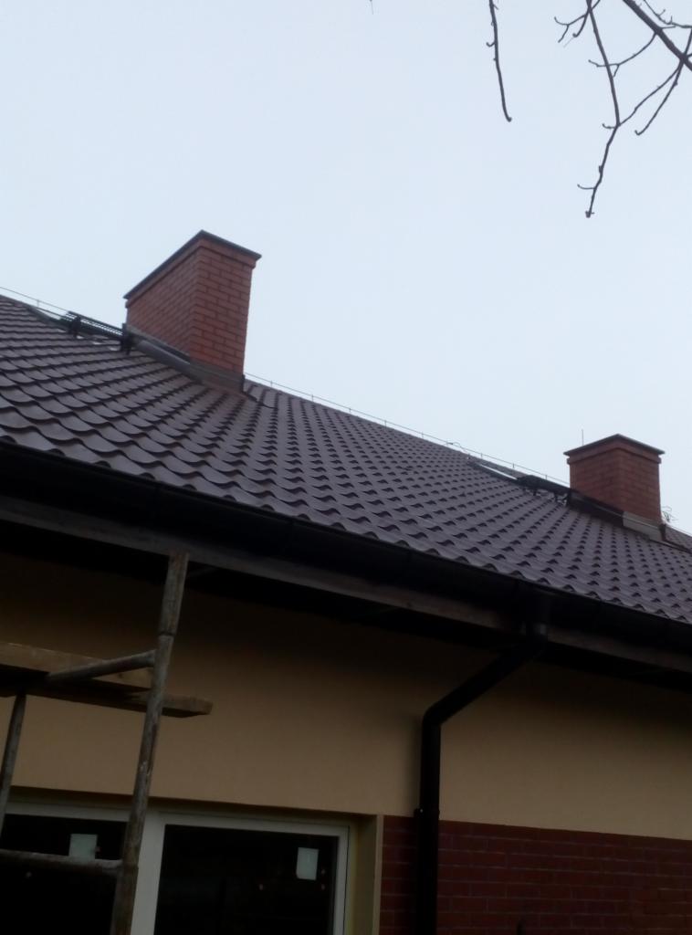 Ciemnobrązowy dach z blachodachówki z dwoma ceglanymi kominami, widok z dołu, fragment elewacji budynku i drabina dekarska z boku.