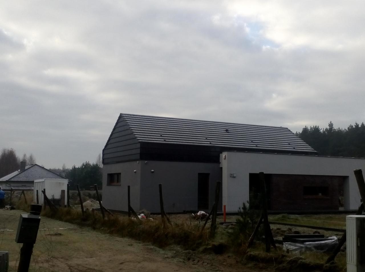 Nowoczesny dom jednorodzinny z ciemną dachówką ceramiczną w pochmurny dzień, widoczny fragment ogrodzenia i sąsiedni budynek.