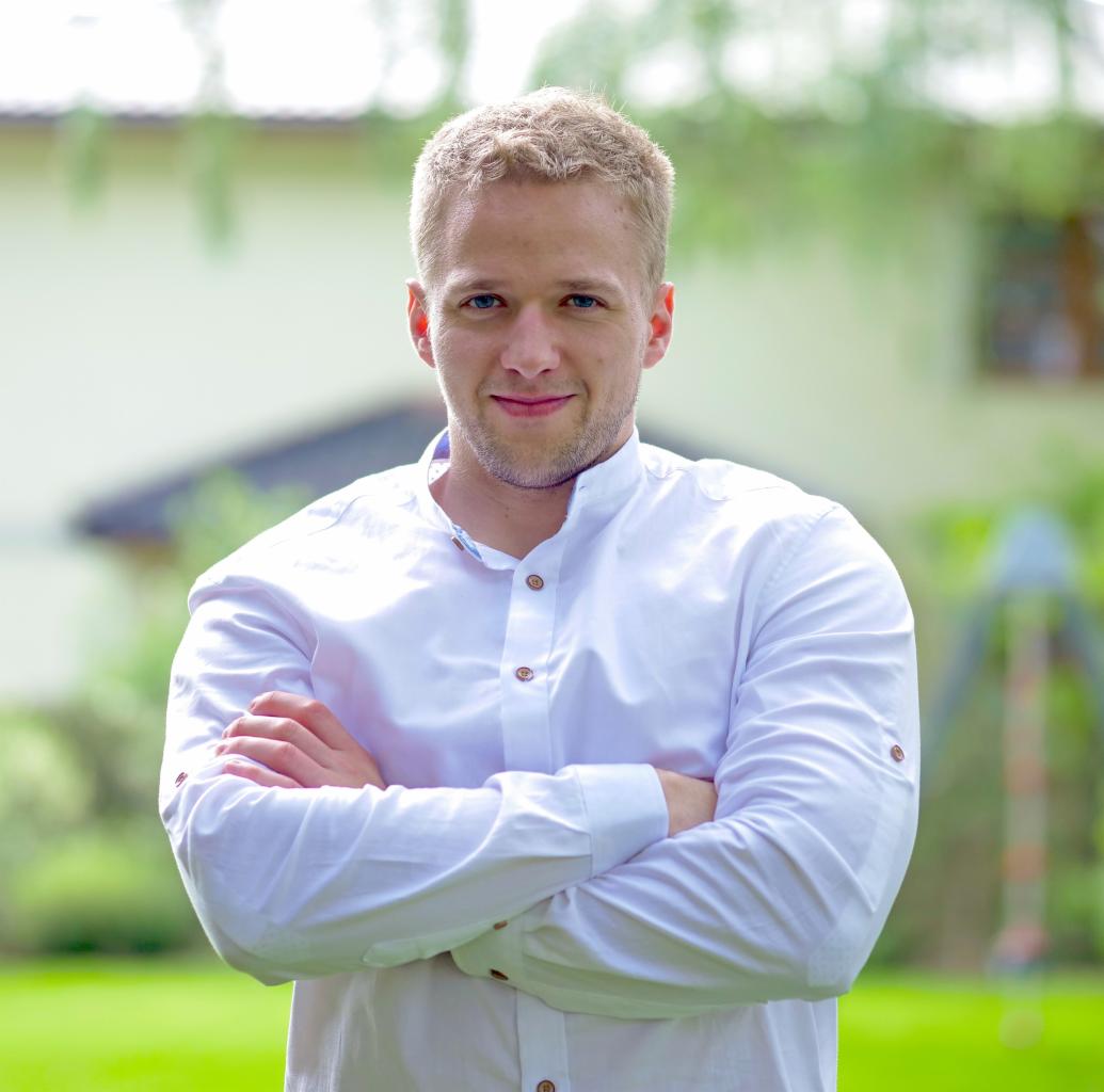 Portret uśmiechniętego blondyna w białej koszuli, skrzyżowane ręce na tle zieleni i jasnego budynku. Delikatne światło dzienne.