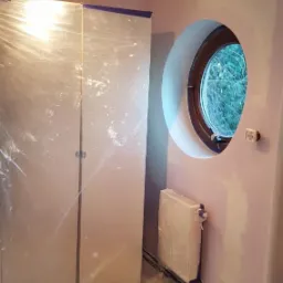 Pokój przygotowany do malowania: meble i podłoga zabezpieczone folią, ściany częściowo pomalowane na jasny róż, okrągłe okno z drewnianą ramą, widoczny grzejnik i lampa sufitowa.