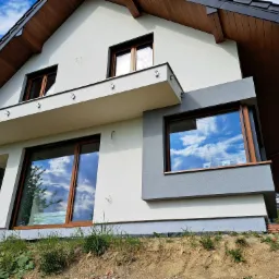 Nowoczesny dom z jasną elewacją i drewnianymi elementami, okna z odbiciem nieba, widok z dołu, podkreślający bryłę budynku i użyte materiały wykończeniowe.