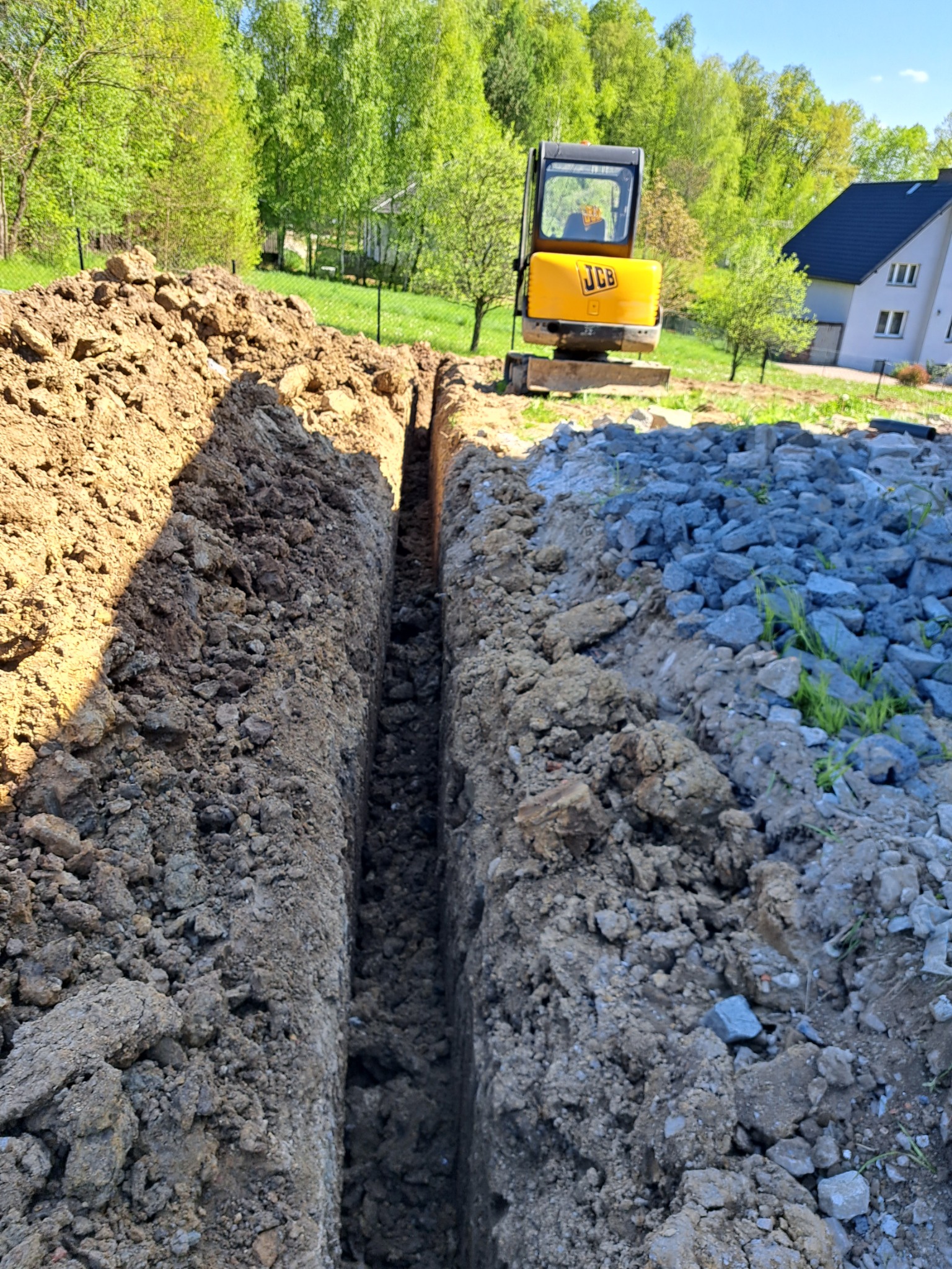Wykopany wąski rów w ziemi, obok sterta gruzu i mała żółta koparka JCB na tle domu i drzew.