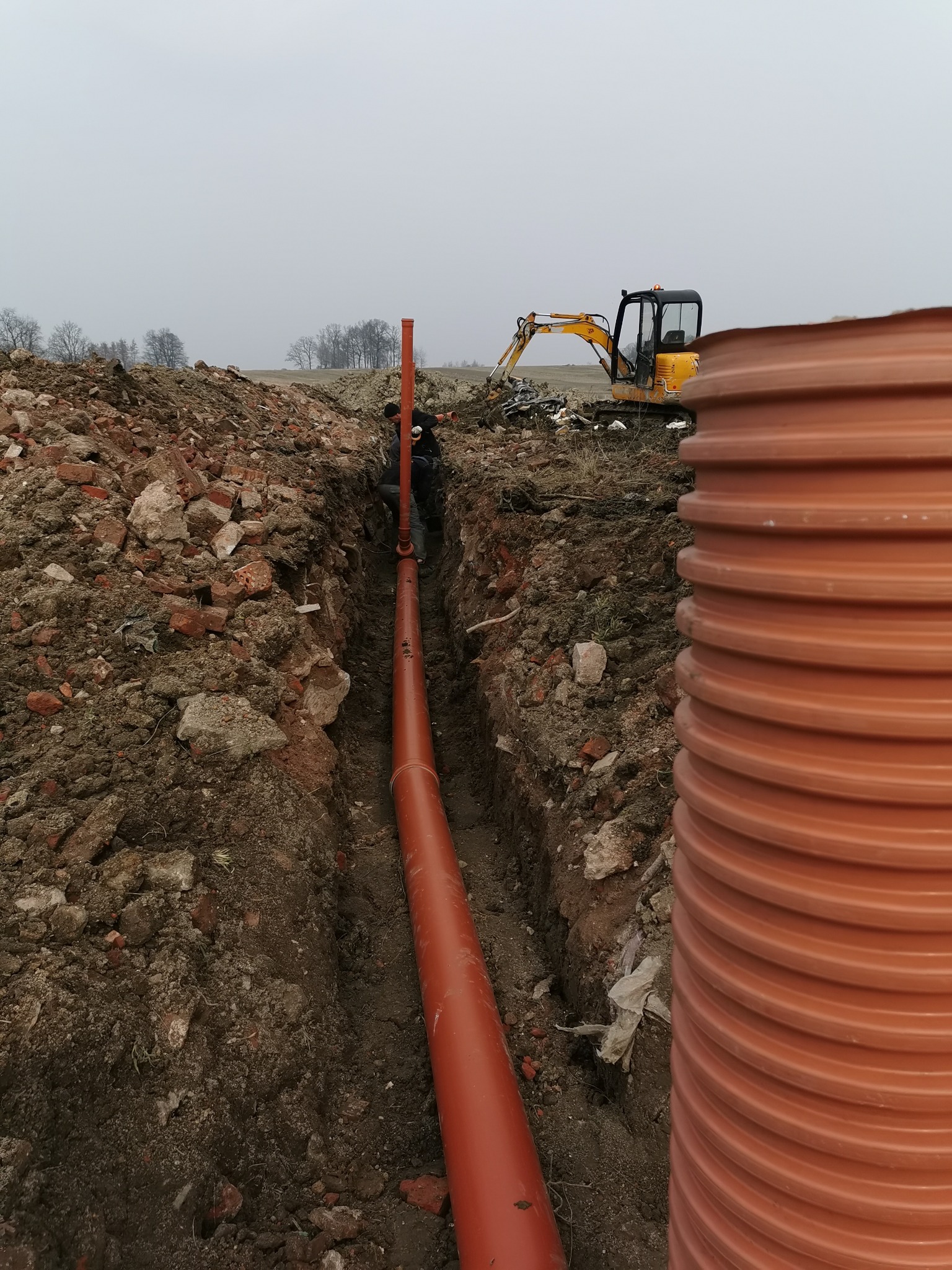 Montaż pomarańczowej rury kanalizacyjnej w wykopie, widoczny pracownik, mini koparka i stos rur w tle.