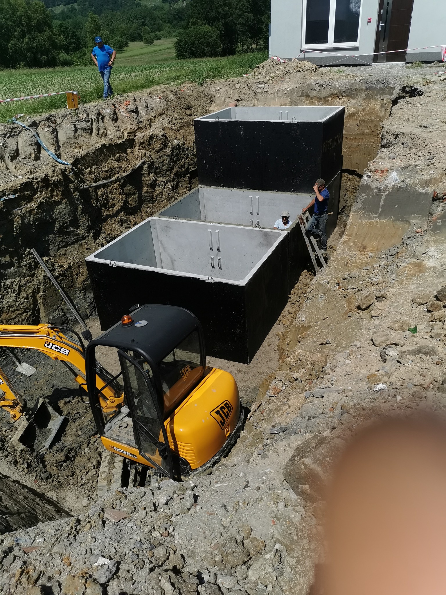 Montaż betonowego szamba dwukomorowego w wykopie przy użyciu mini koparki JCB, widoczni dwaj pracownicy na drabinie i na górze wykopu.