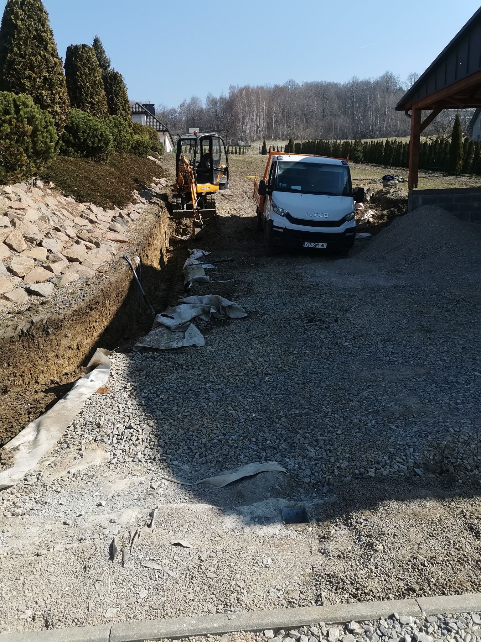 Mini koparka i samochód ciężarowy podczas prac ziemnych na terenie posesji, widoczny wykop, kamień i materiał geowłókninowy.