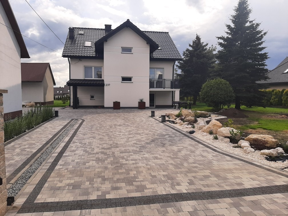Dwukondygnacyjny dom z jasną elewacją, czarnym dachem i numerem 237, otoczony posesją z nowo ułożoną kostką brukową, ozdobnymi kamieniami i zielenią.