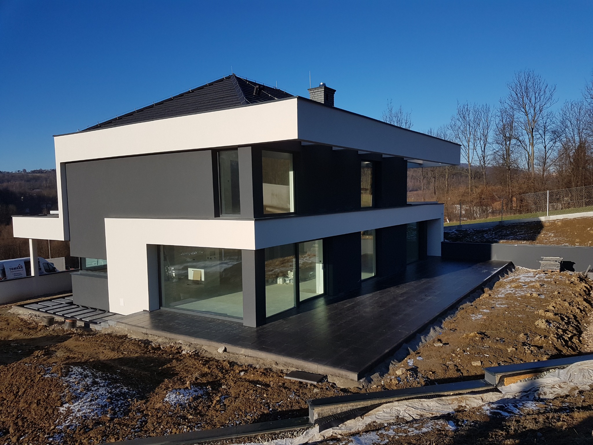 Nowoczesny dwukondygnacyjny dom z płaskim dachem, wykończony w bieli i szarości, z dużymi przeszkleniami, otoczony ziemią w trakcie prac wykończeniowych.