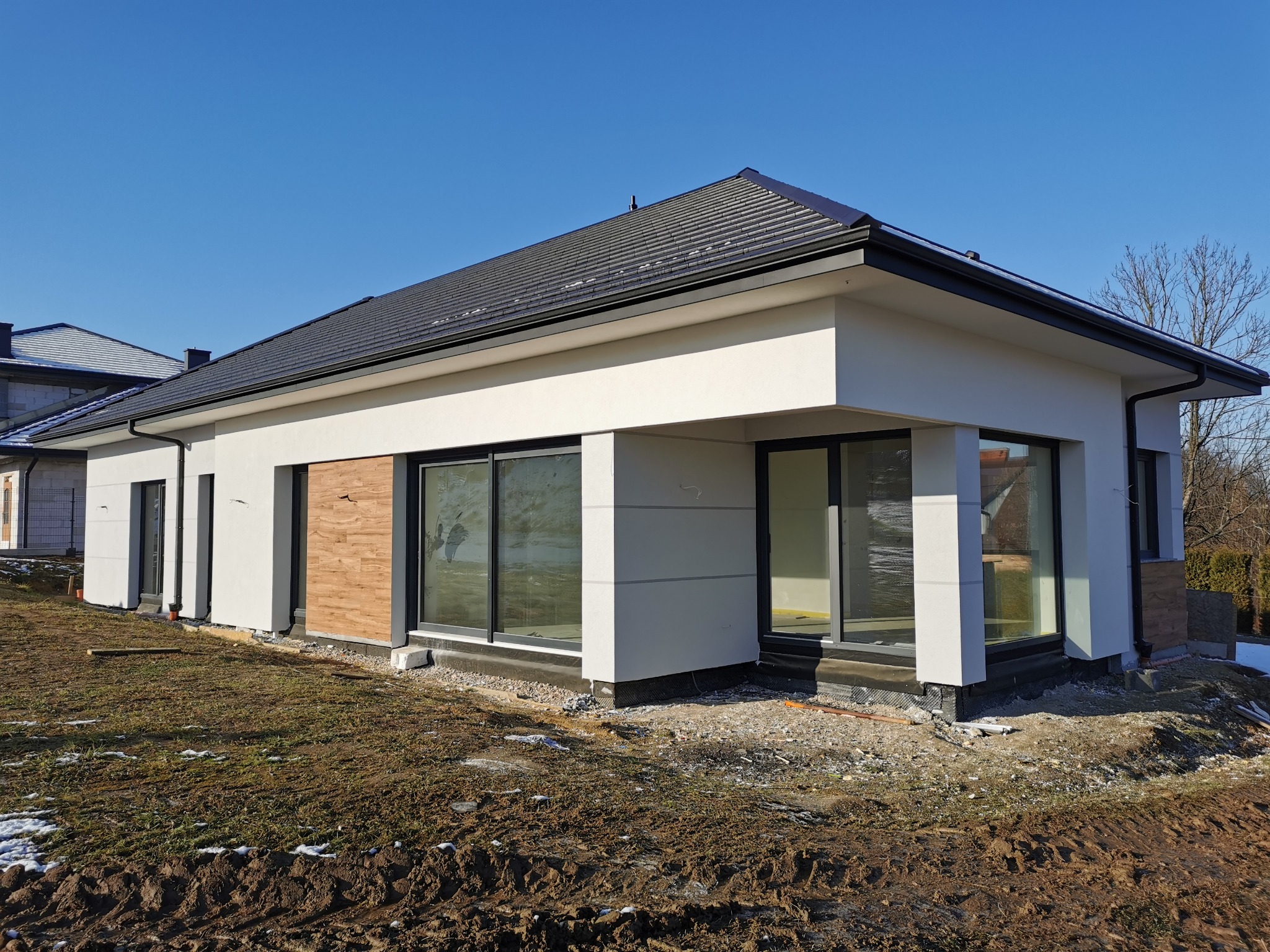 Nowoczesny dom jednorodzinny w Radziszowie z elementami dekoracyjnymi z drewna na elewacji, duże okna, ciemna dachówka, teren wokół domu w trakcie zagospodarowania.