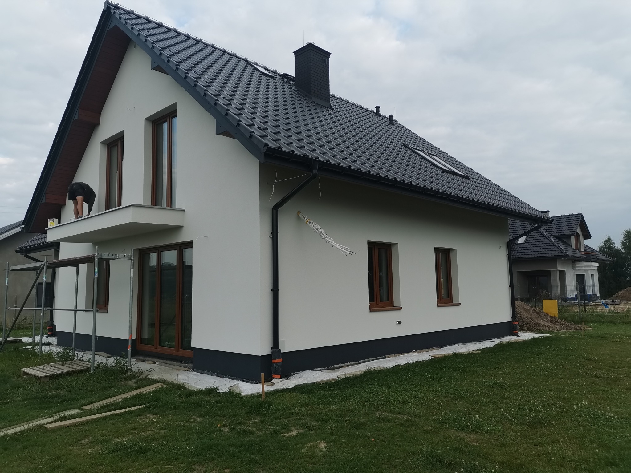 Wykończenie elewacji domu jednorodzinnego z ciemną dachówką, widoczne rusztowanie i pracownik na balkonie, świeżo pomalowana ściana z brązowymi oknami.