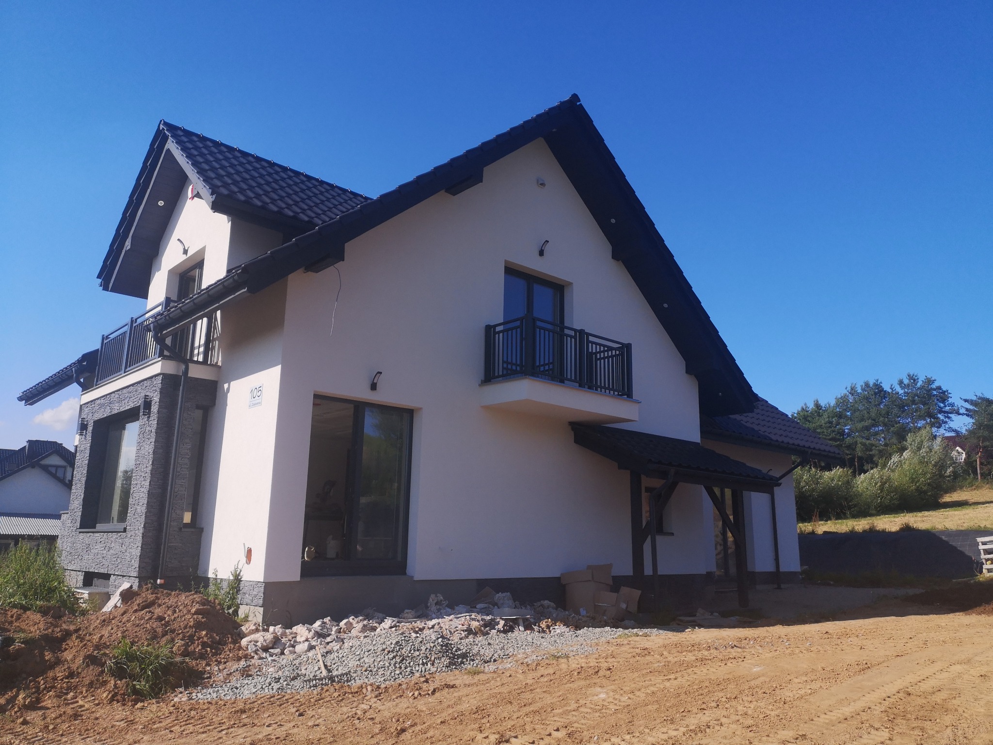 Nowoczesny dom jednorodzinny z białymi ścianami i czarnym dachem, z balkonem z metalową balustradą, widok z zewnątrz na tle błękitnego nieba, teren wokół domu w trakcie zagospodarowywania.
