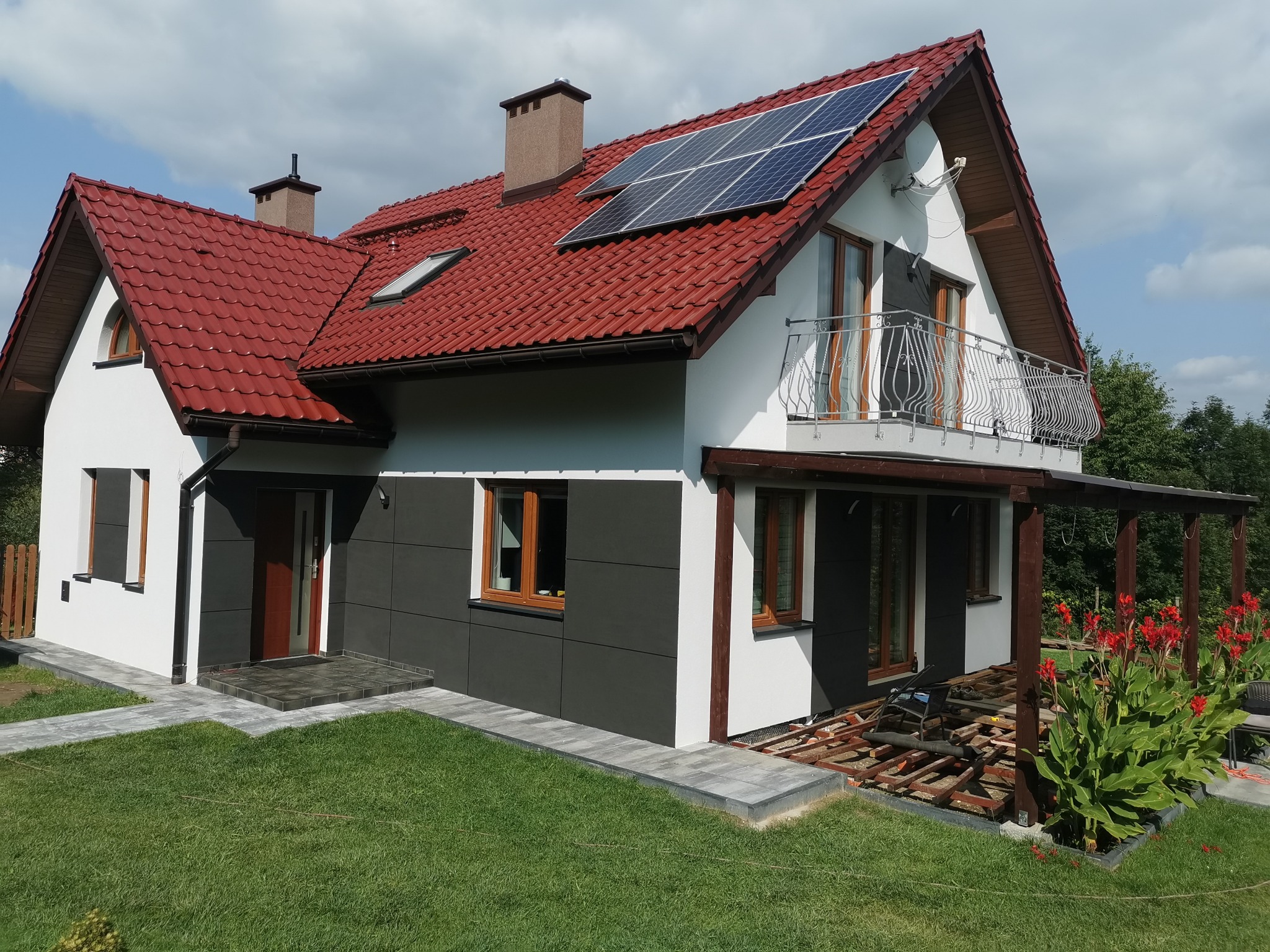 Nowoczesny dom z panelami słonecznymi na czerwonym dachu, białą i szarą elewacją, oraz zadaszeniem tarasu w trakcie budowy.