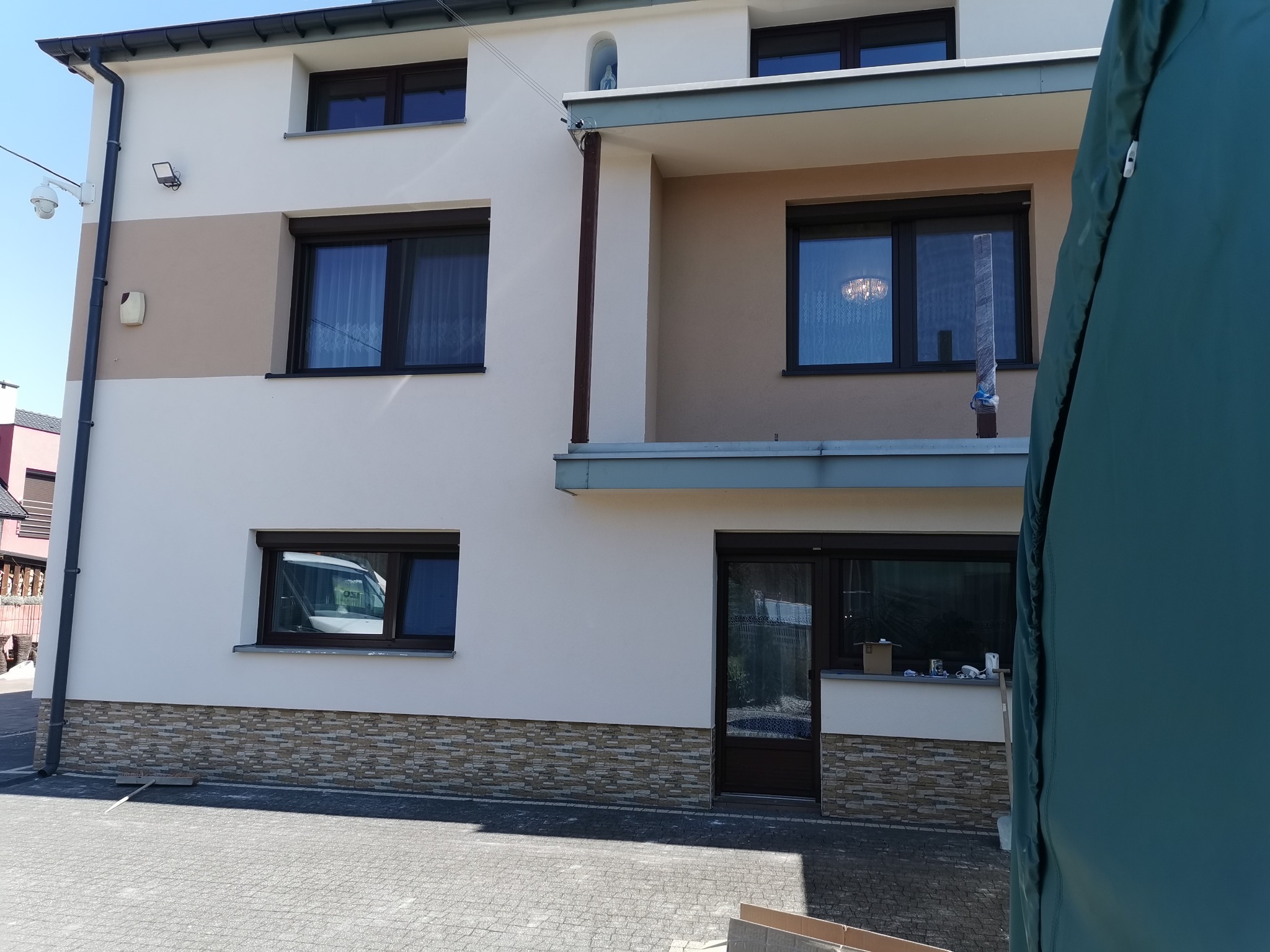 Dwukondygnacyjny dom z jasną elewacją, brązowymi oknami i dekoracyjnym kamieniem na fundamencie, widoczny z zewnątrz, z fragmentem zielonej plandeki w kadrze.