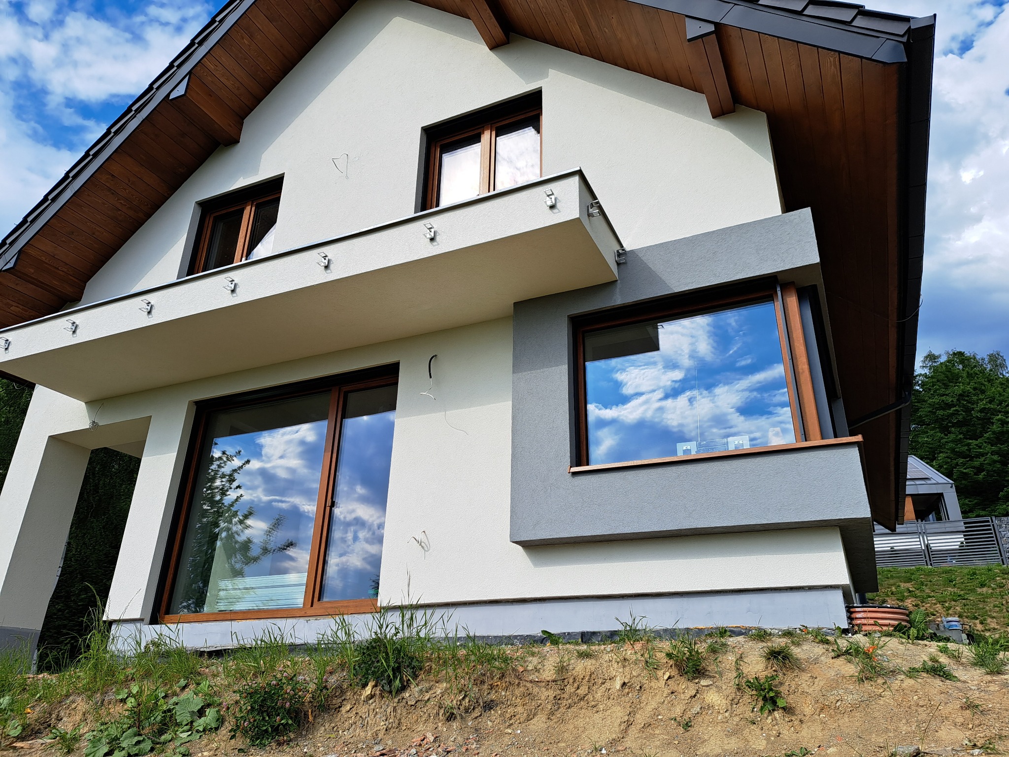 Nowoczesny dom z jasną elewacją i drewnianymi elementami, okna z odbiciem nieba, widok z dołu, podkreślający bryłę budynku i użyte materiały wykończeniowe.