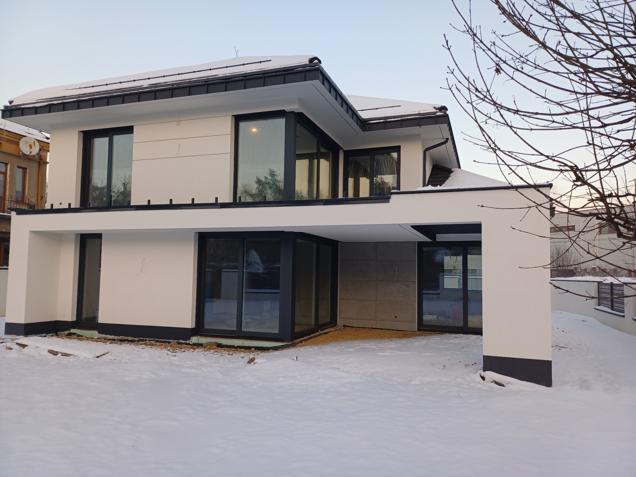 Nowoczesny dom z płaskim dachem w zimowej scenerii. Biała elewacja kontrastuje z czarnymi ramami okien. Widoczny śnieg i fragmenty ocieplenia przy fundamentach.