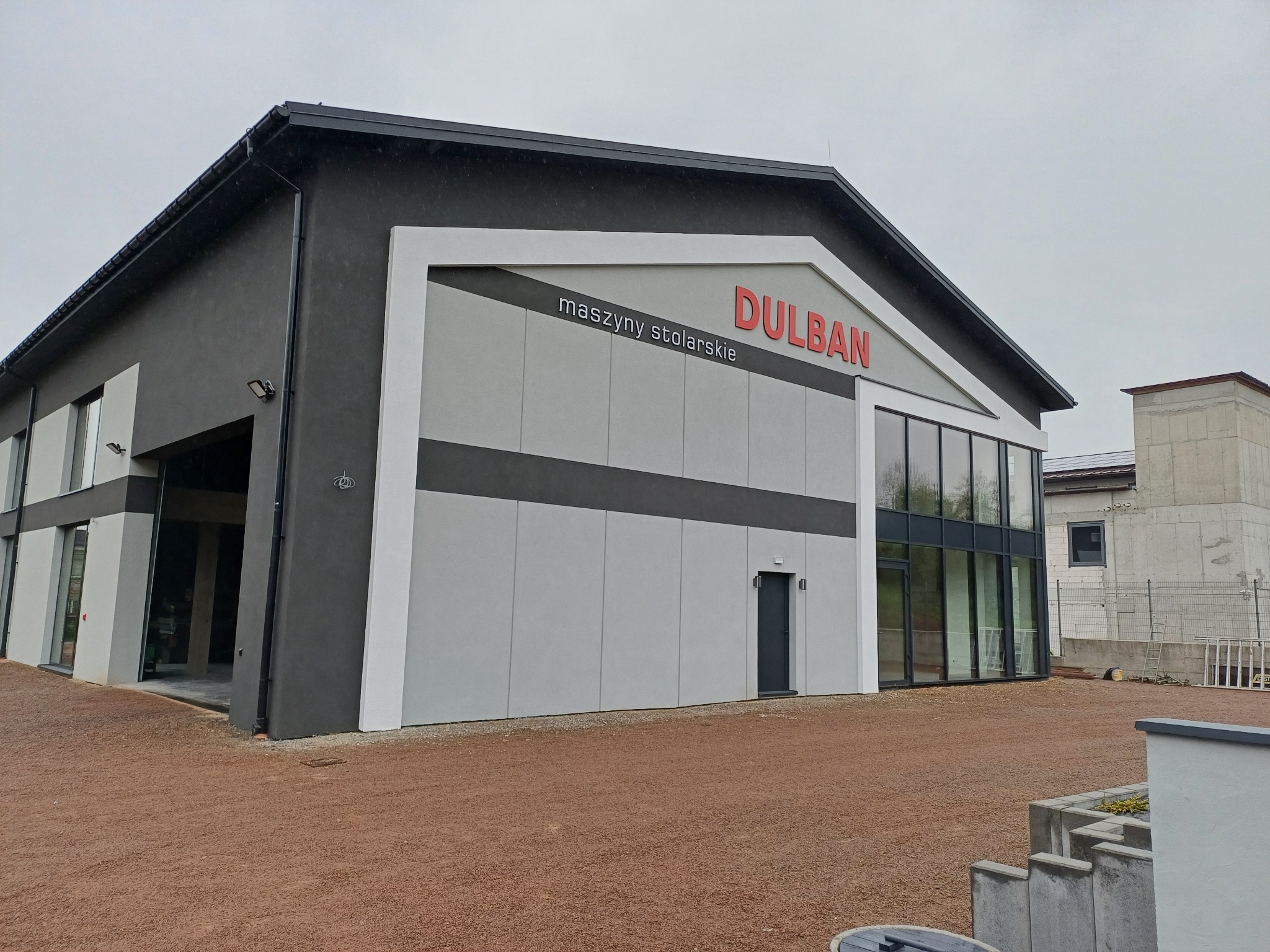 Nowoczesny budynek z szarą elewacją, dużymi oknami i czerwonym logo 'DULBAN maszyny stolarskie' na tle pochmurnego nieba. Widoczny plac z utwardzoną nawierzchnią.