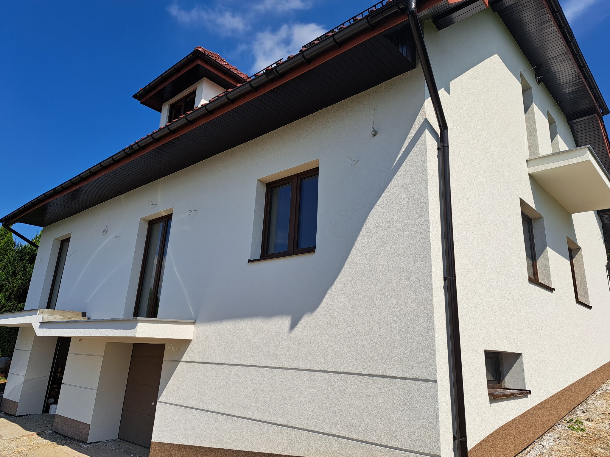 Dwukondygnacyjny dom z jasną elewacją o strukturze baranka, brązowymi oknami i dachem, widoczny z dołu pod kątem, w słoneczny dzień.