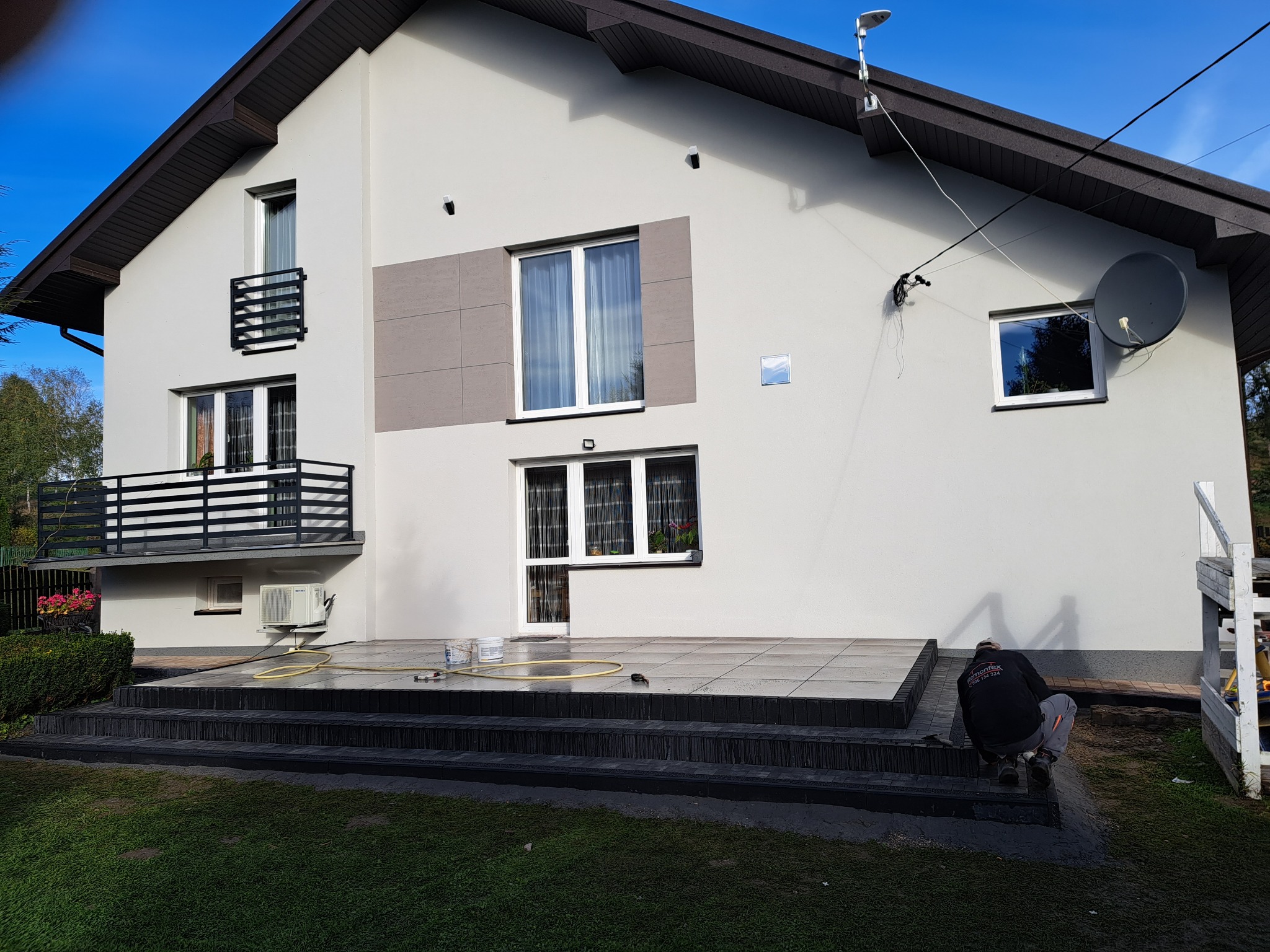 Elewacja domu po tynkowaniu i malowaniu. Widoczny pracownik wykonujący prace wykończeniowe na tarasie z kostki. Dom w odcieniach szarości z czarnymi detalami.