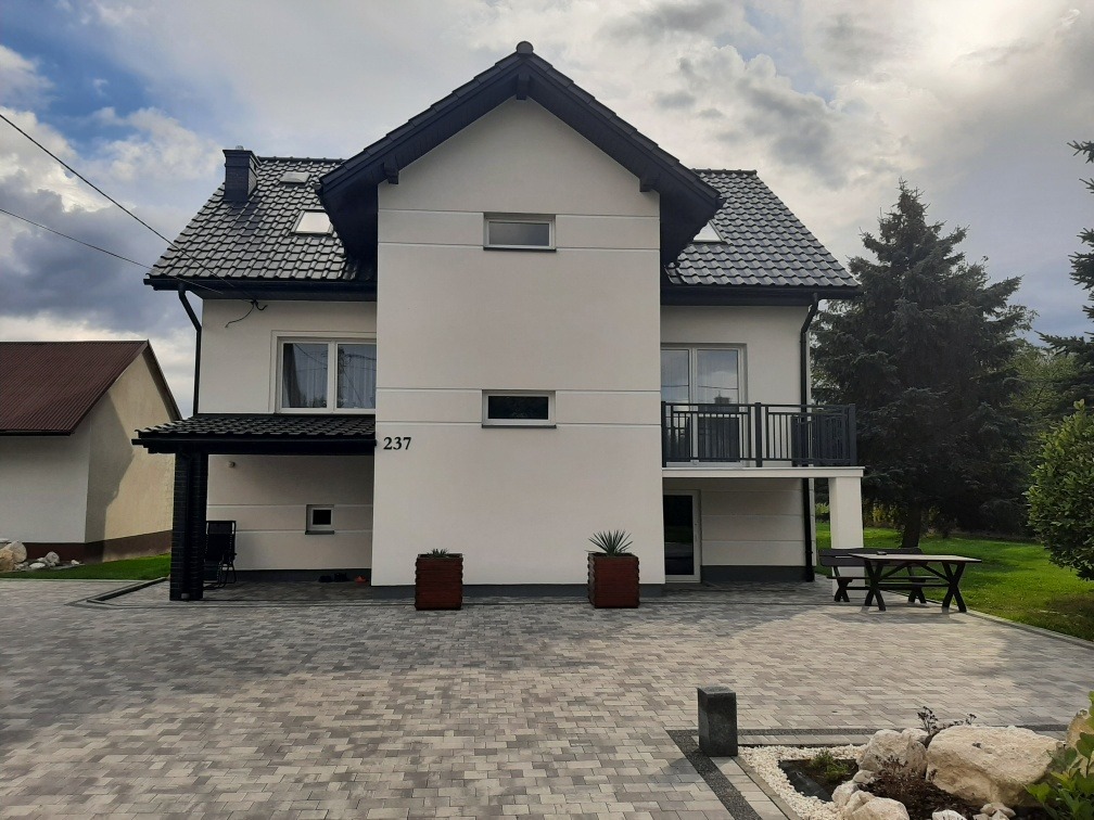 Dwukondygnacyjny dom z białymi ścianami i ciemnym dachem, numer 237, otoczony kostką brukową i zielenią. Widoczne elementy dekoracyjne: donice z roślinami, lampy.