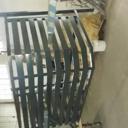 Metalowa konstrukcja balustrady po nałożeniu czarnej powłoki, zawieszona w warsztacie, widoczna pod światło okna.