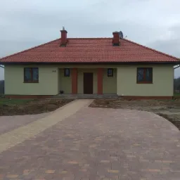 Nowo wyłożony, zakrzywiony podjazd z dwukolorowej kostki prowadzący do parterowego domu z czerwoną dachówką w pochmurny dzień. Widoczny numer domu 340.