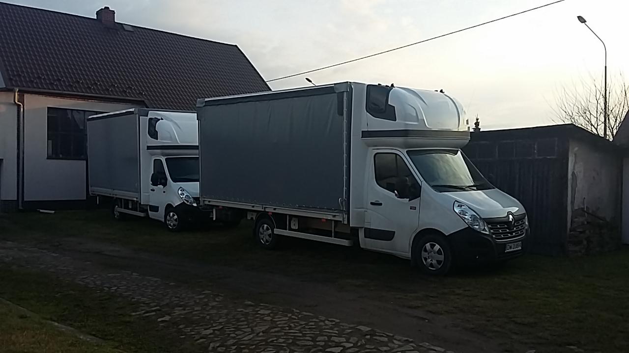 Dwa białe busy transportowe z szarą plandeką zaparkowane na posesji przed domem i garażem, widoczne z boku w pochmurny dzień.