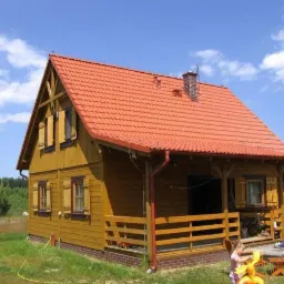 Niski, parterowy dom z bali, z czerwonym dachem i drewnianymi okiennicami, otoczony zieloną trawą, z dzieckiem bawiącym się na tarasie i osobą pracującą w oddali.