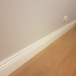 mario-dario - Nowa, biała listwa przypodłogowa z dekoracyjnym frezem, zamontowana przy jasnej ścianie i drewnianej podłodze, widoczny fragment gniazdka elektrycznego.