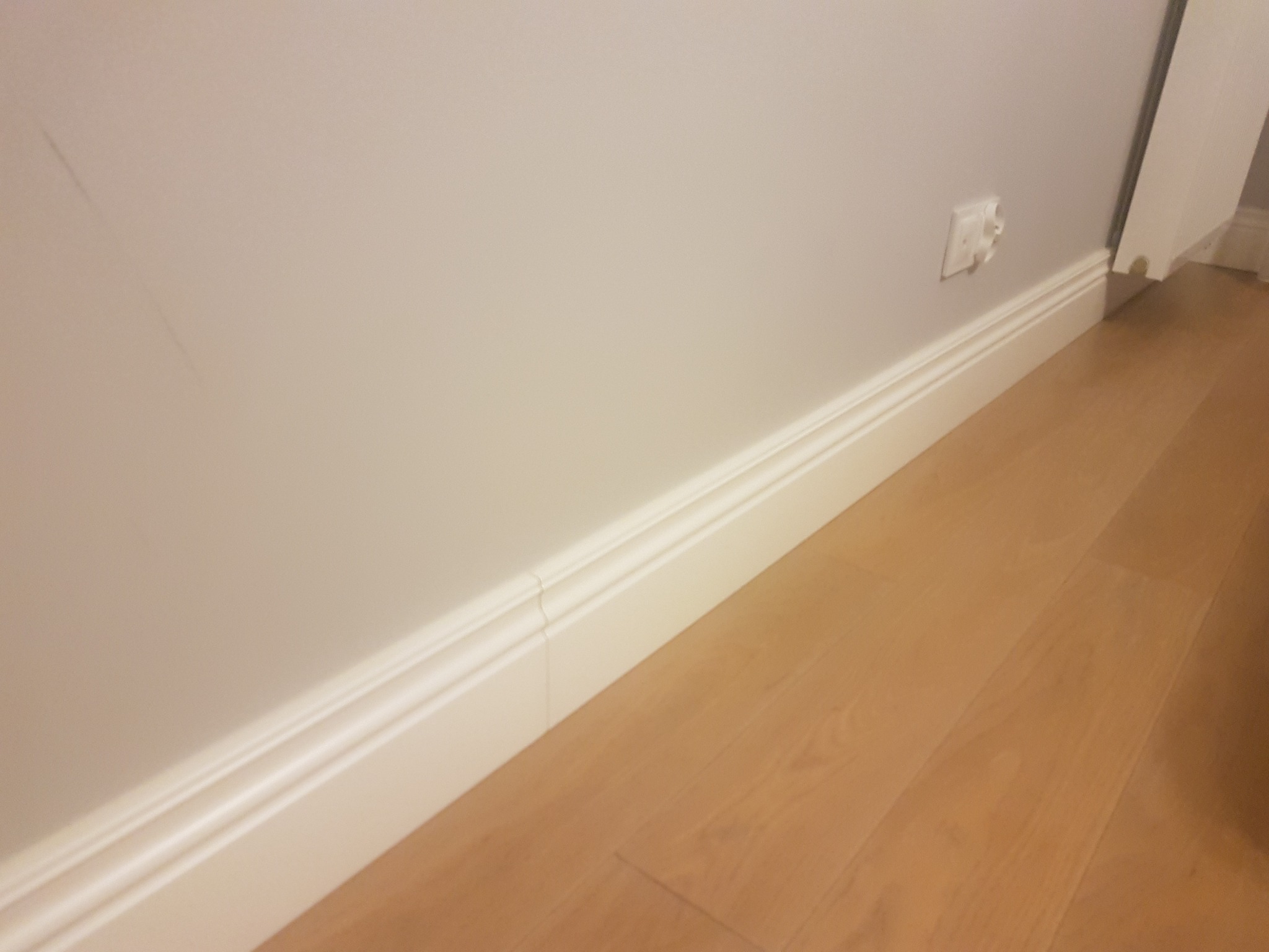 Nowa, biała listwa przypodłogowa z dekoracyjnym frezem, zamontowana przy jasnej ścianie i drewnianej podłodze, widoczny fragment gniazdka elektrycznego.