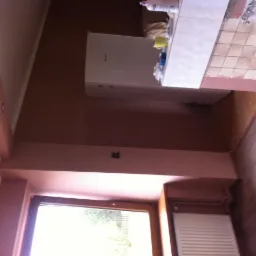 Ściana w odcieniach brązu, świeżo pomalowana, z widocznym oknem i kaloryferem; w tle fragment łazienki z płytkami i umywalką.