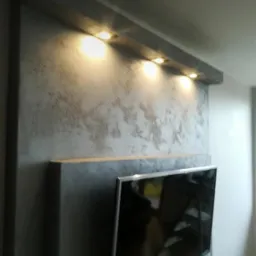 Szara ściana z dekoracyjną fakturą, oświetlona punktowymi lampami LED, z fragmentem wiszącego telewizora i nakładką ekranu smartfona z informacją o połączeniu przychodzącym.