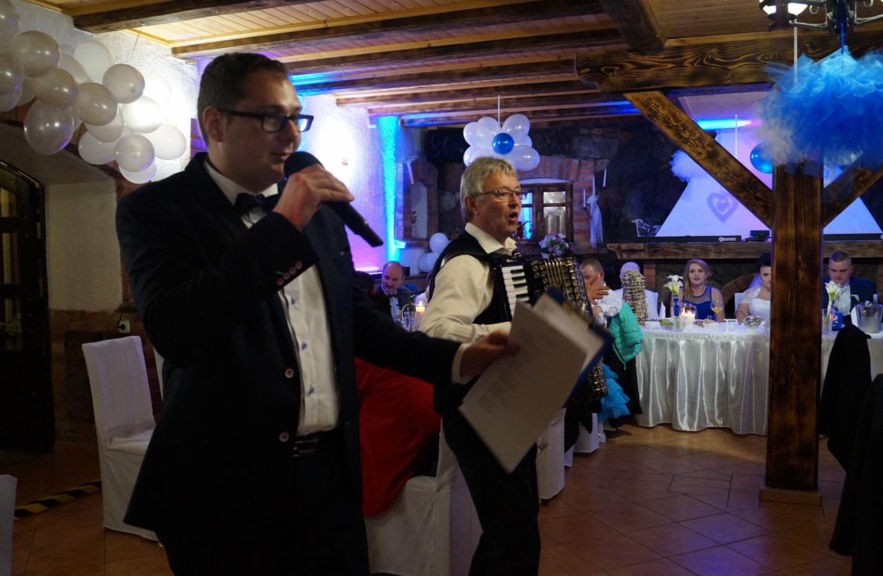 Mężczyzna w garniturze z mikrofonem prowadzi uroczystość, obok akordeonista z kartką, w tle udekorowana sala weselna z białymi balonami i niebieskim oświetleniem.