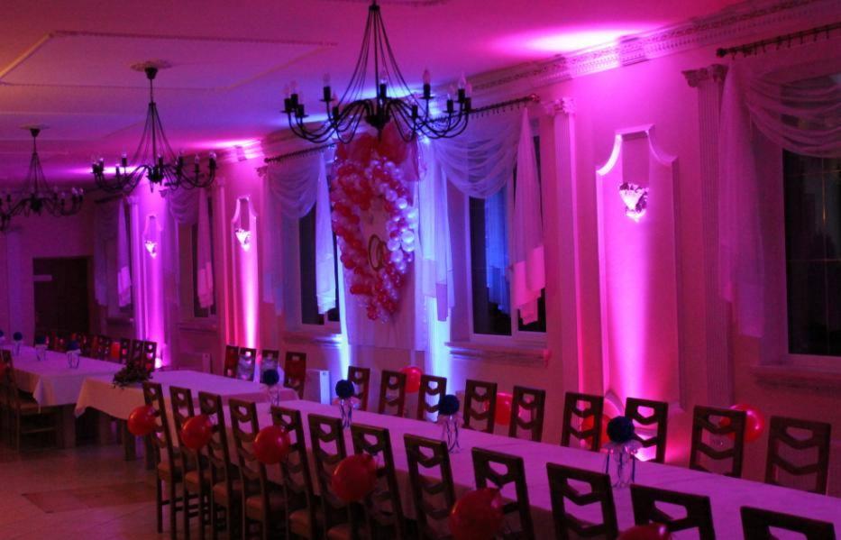 Sala bankietowa z długimi stołami udekorowanymi balonami, oświetlona fioletowym i niebieskim światłem, z dekoracją balonową na ścianie.
