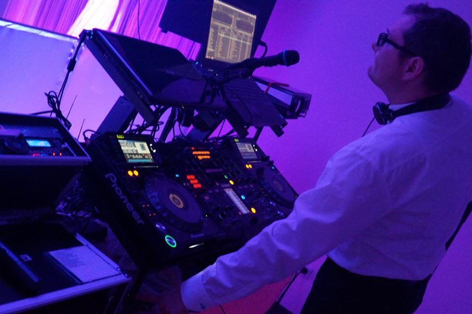 DJ w białej koszuli obsługuje profesjonalny sprzęt Pioneer podczas imprezy, oświetlony fioletowym światłem, z mikrofonem i słuchawkami na szyi.