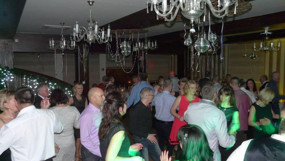 Uczestnicy imprezy tanecznej pod dekoracyjnymi żyrandolami i dyskotekowymi kulami, widoczna część schodów z lampkami.