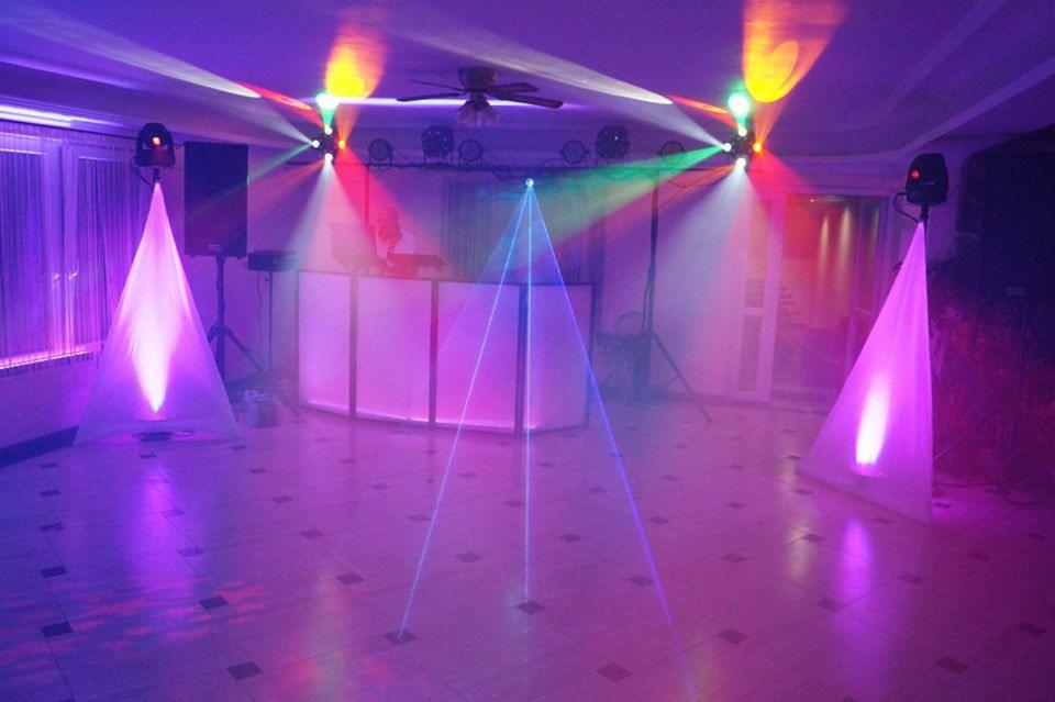 Sala z oświetleniem eventowym: reflektory na statywach z białymi abażurami, kolorowe światła i laserowe wiązki, stanowisko DJ-a w tle.