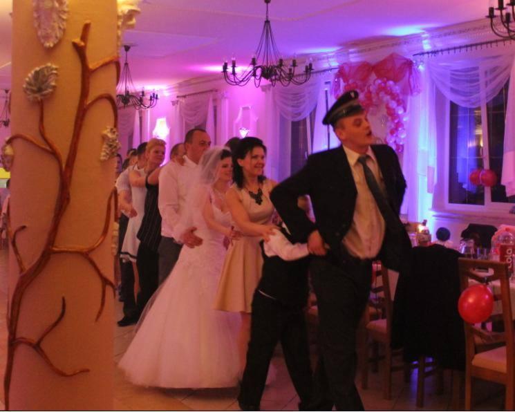 Goście weselni w trakcie korowodu tanecznego prowadzonego przez wodzireja w czapce, za parą młodą, w sali z dekoracją światłem i balonami.