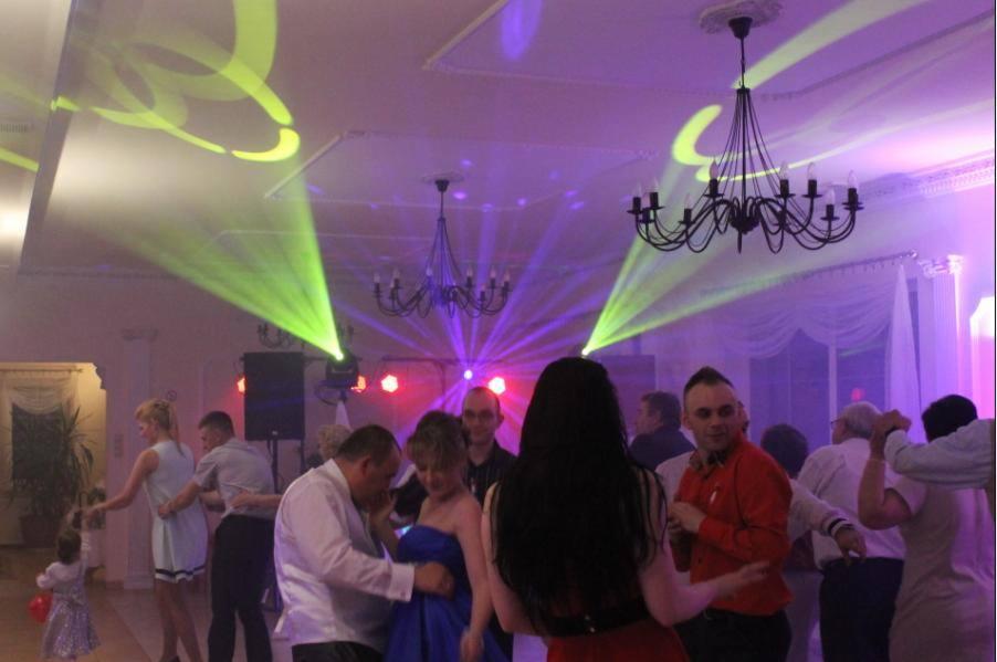 Parkiet taneczny z bawiącymi się gośćmi, oświetlony zielonymi i fioletowymi światłami reflektorów, z czarnymi żyrandolami na suficie i dekoracjami.