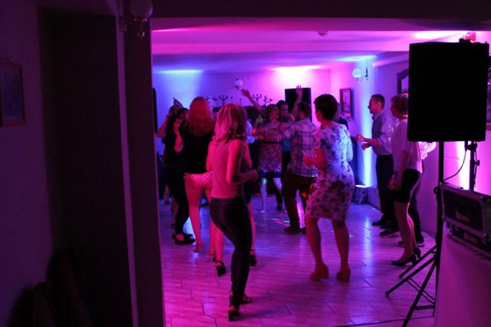 Tańczący ludzie na parkiecie podświetlonym fioletowymi i niebieskimi światłami, widoczny sprzęt nagłośnieniowy.