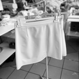 MINU Anna Pabisiak - Czarno-białe zdjęcie białej spódniczki na wieszaku w pracowni krawieckiej, z maszynami do szycia i pracownikami w tle.