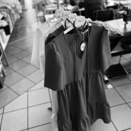 MINU Anna Pabisiak - Dwa gotowe do wysyłki, damskie, letnie sukienki na wieszakach, z metkami producenta, w tle szwalnia z pracującymi osobami.