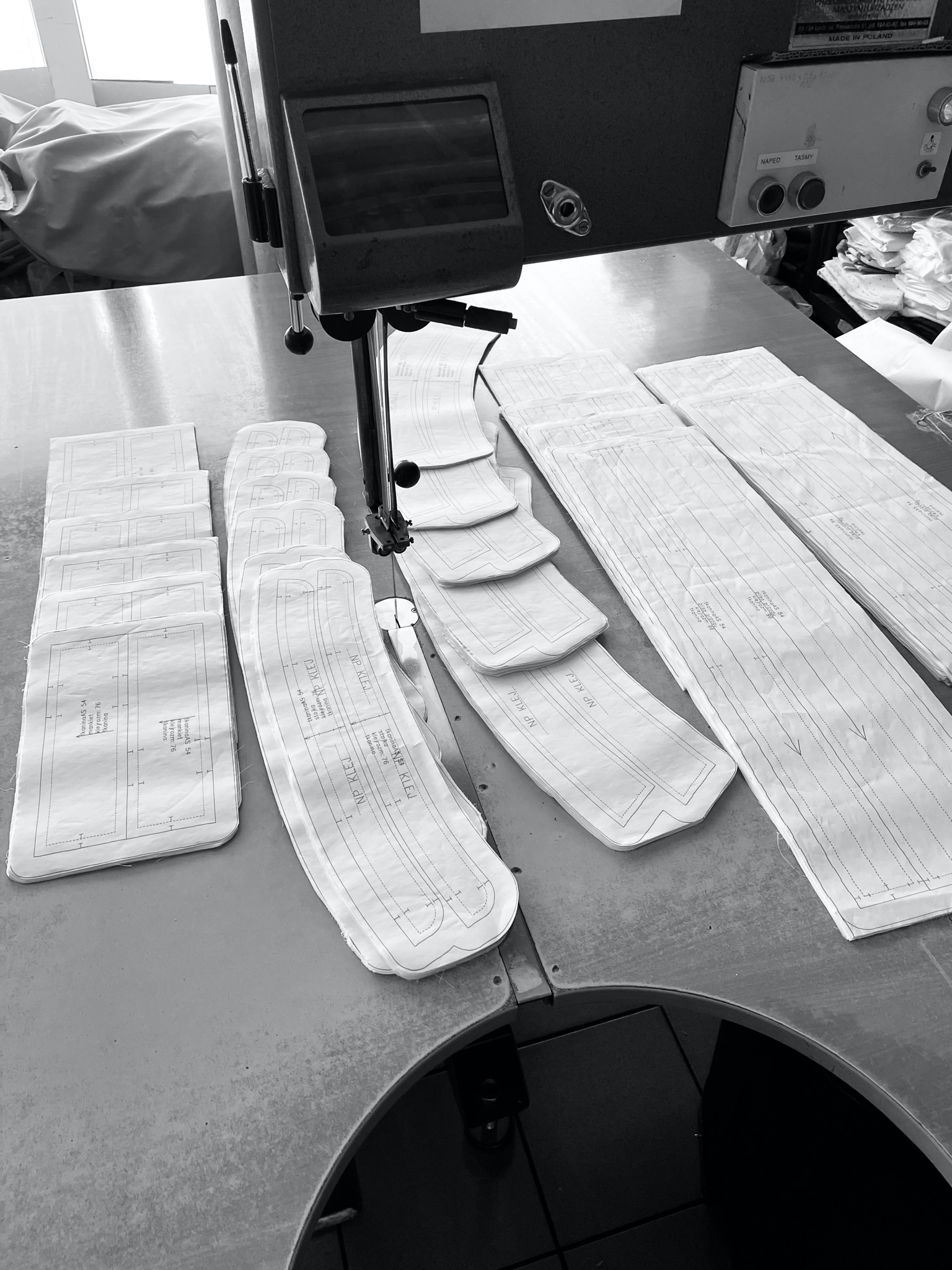 Czarno-białe zdjęcie przedstawia wykroje odzieżowe ułożone na stole pod maszyną krojczą. W tle widoczne stosy materiałów i panel sterowania maszyny z przyciskami 'NAPĘD' i 'TAŚMY' oraz napisem...
