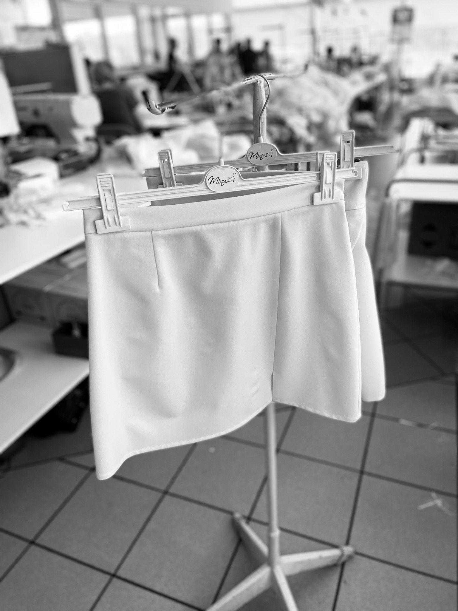 Czarno-białe zdjęcie białej spódniczki na wieszaku w pracowni krawieckiej, z maszynami do szycia i pracownikami w tle.
