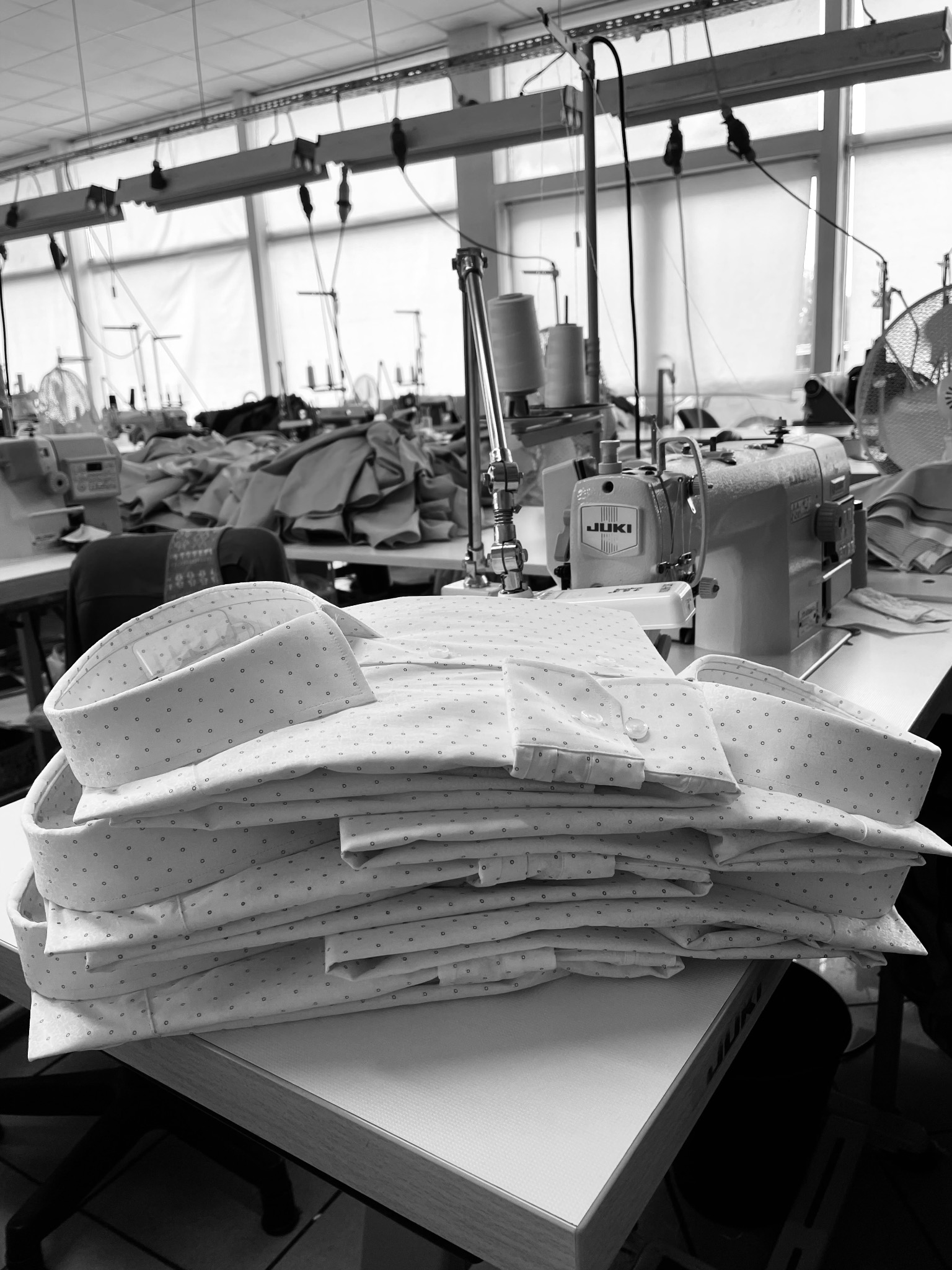 Stos starannie złożonych, jasnych koszul męskich w drobne wzory, na tle warsztatu z maszynami do szycia, w czerni i bieli.