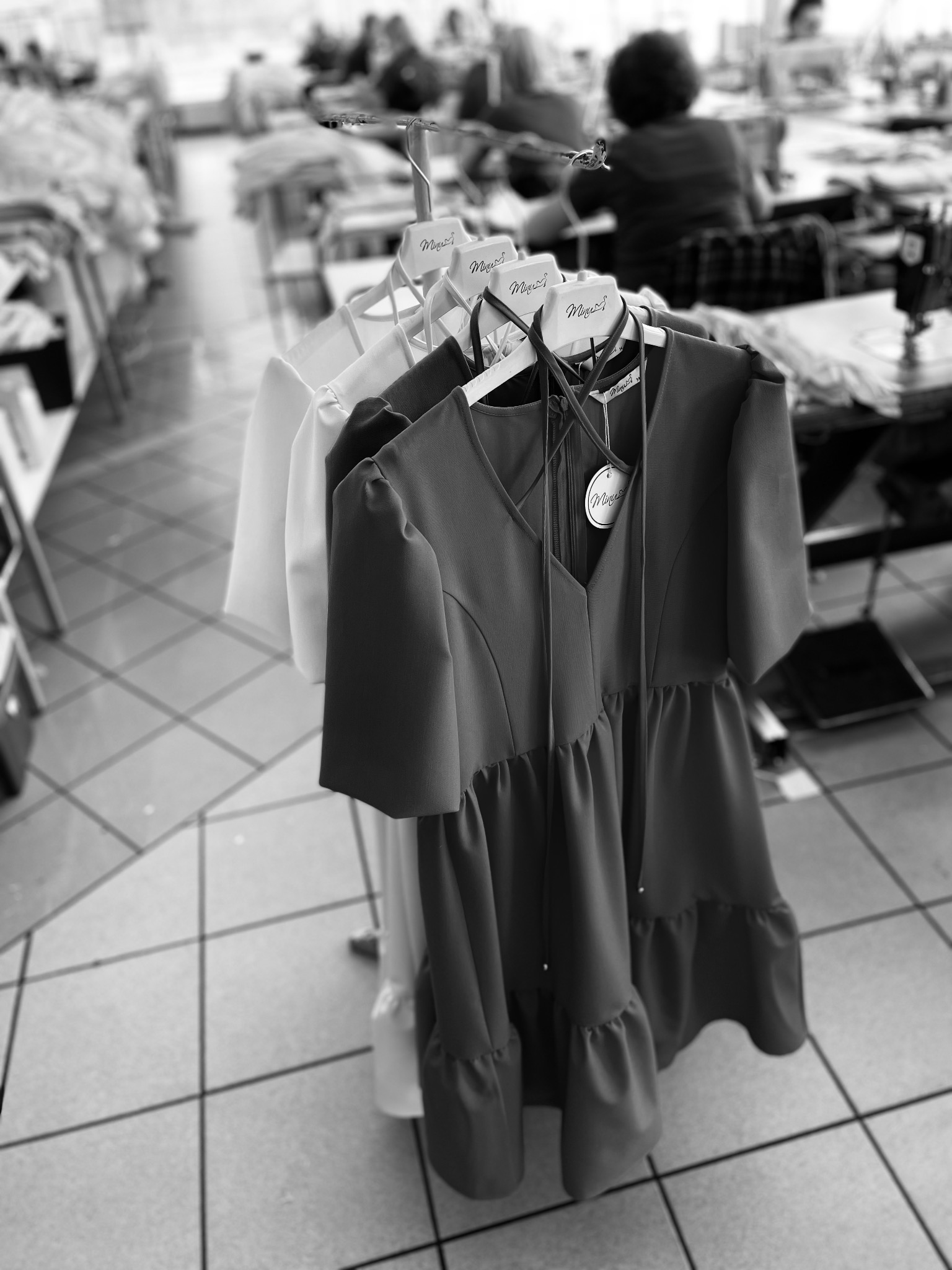 Dwa gotowe do wysyłki, damskie, letnie sukienki na wieszakach, z metkami producenta, w tle szwalnia z pracującymi osobami.